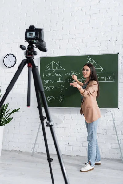 Profesor escribiendo fórmula matemática en pizarra cerca de la cámara digital en clase - foto de stock