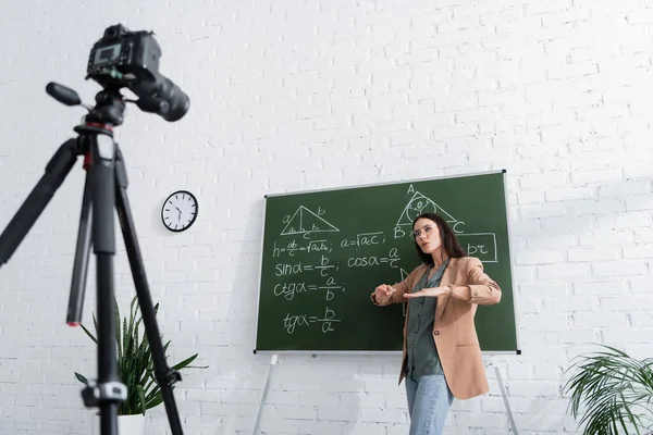 Maestro gesticulando cerca de pizarra con fórmulas matemáticas y cámara digital en el aula — Stock Photo