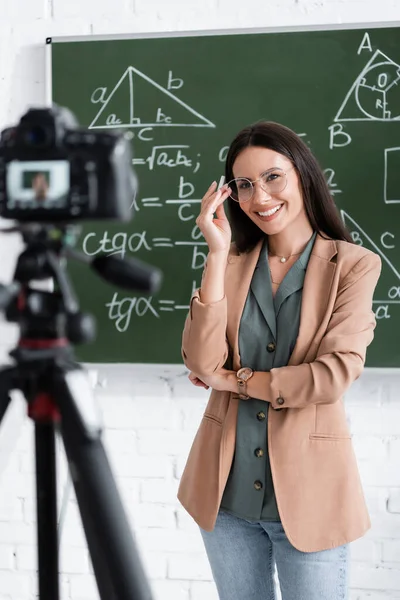 Cheerful teacher holding chalk near chalkboard and digital camera in classroom — Foto stock