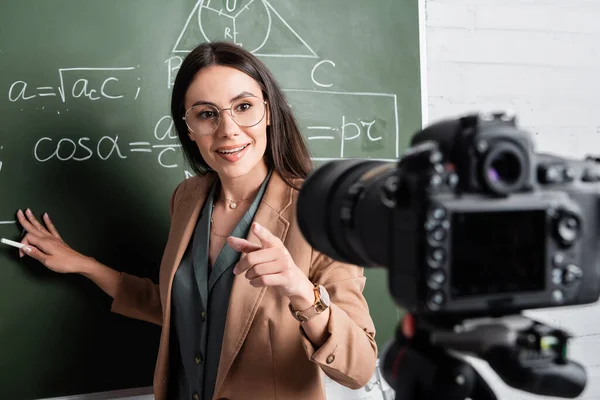 Smiling teacher holding chalk near chalkboard with formulas and digital camera — Foto stock