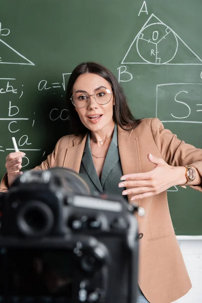 Profesor hablando cerca de fórmulas matemáticas en pizarra y cámara digital borrosa en el aula - foto de stock