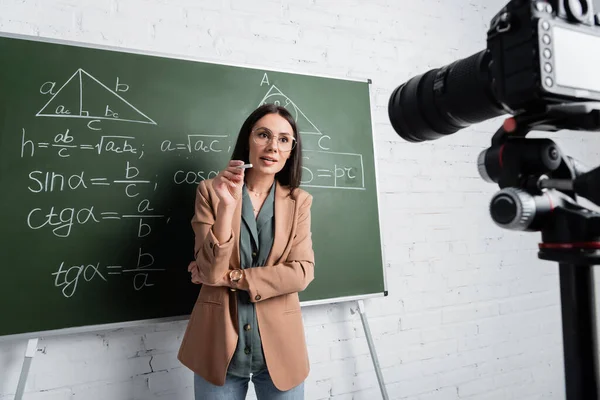 Teacher holding chalk and talking near chalkboard with formulas and digital camera — Foto stock