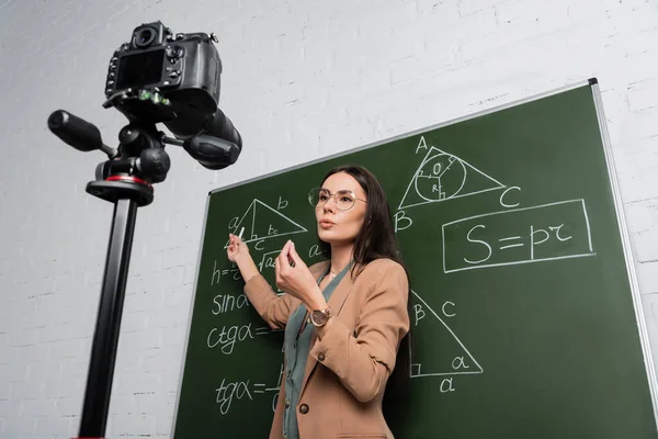 Niedrigwinkel-Ansicht eines Lehrers, der in der Schule in der Nähe von Mathematikformeln auf Tafel und Digitalkamera spricht — Stockfoto