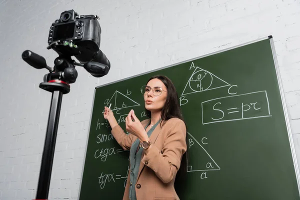 Vista de bajo ángulo del profesor hablando cerca de fórmulas matemáticas en pizarra y cámara digital en la escuela - foto de stock