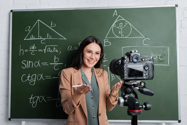 Positive teacher pointing at digital camera near chalkboard with math formulas — Stock Photo