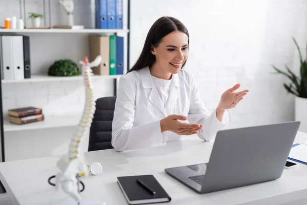 Médico sonriente apuntando a la computadora portátil durante la consulta en línea en el hospital - foto de stock