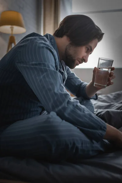 Vue latérale de l'homme en pyjama tenant l'eau tout en souffrant d'insomnie sur le lit — Photo de stock