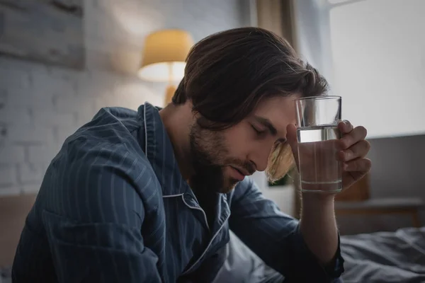 Hombre cansado en pijama sosteniendo un vaso de agua en la cama por la mañana - foto de stock