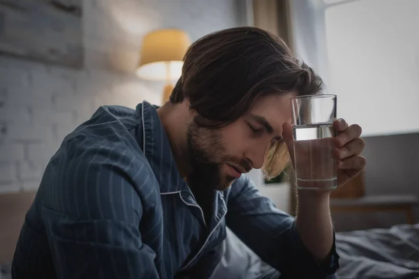 Uomo stanco in pigiama che tiene un bicchiere d'acqua a letto al mattino — Foto stock