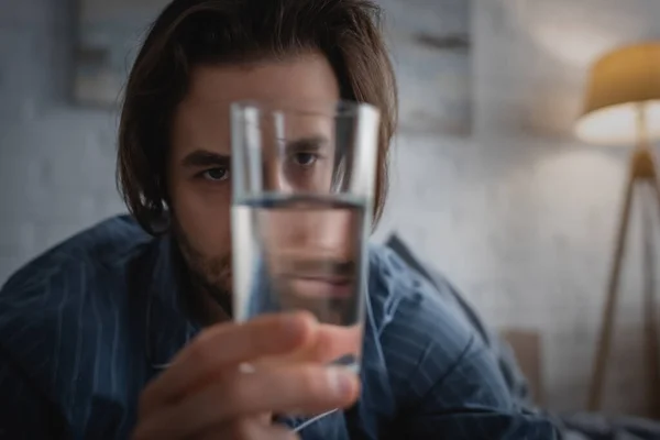 Tired man with sleep disorder holding glass of water at home — Stockfoto