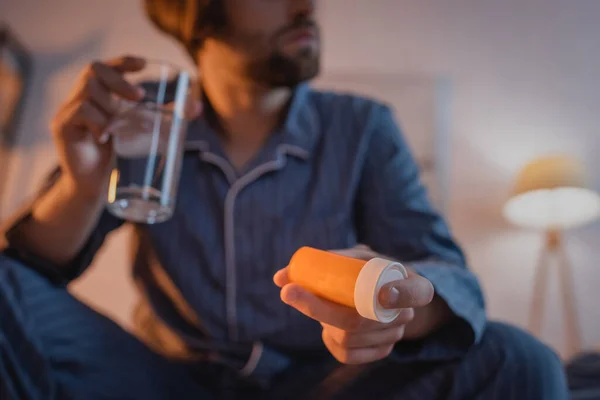 Vue recadrée de l'homme flou en pyjama tenant des pilules et un verre d'eau dans la chambre — Photo de stock