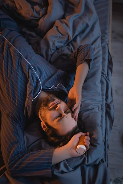 Top view of tired man with insomnia holding pills and touching head on bed — Stock Photo