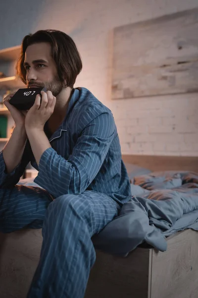 Brunette man in pajama holding alarm clock on bed at night — Stockfoto