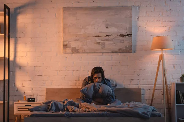 Tired man holding pillow and looking at camera on bed at night — Stock Photo
