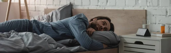 Bearded man looking at alarm clock near water and pills on bedside table in bedroom, banner — Stock Photo