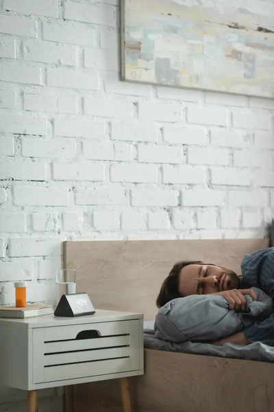 Junger Mann mit geschlossenen Augen liegt auf Kissen in der Nähe von Tabletten und Wecker im Schlafzimmer — Stockfoto