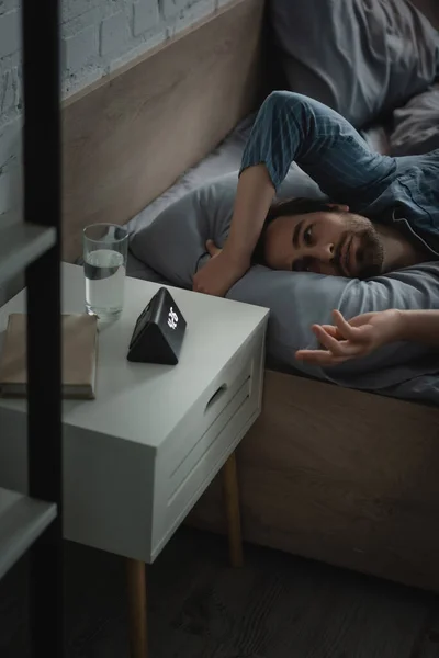 Man with insomnia looking at alarm clock near book and water on bedside table at home - foto de stock