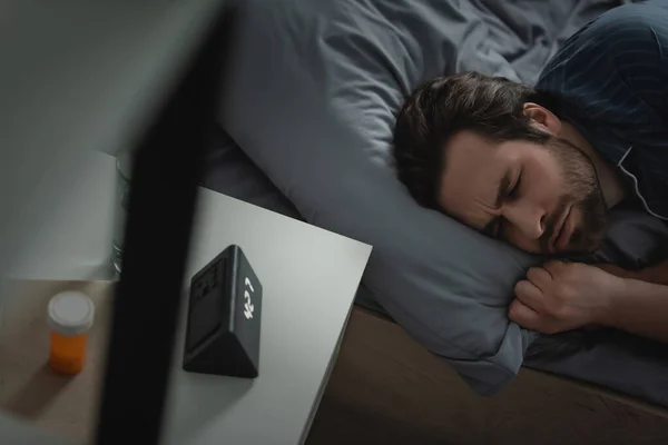 Vista de ángulo alto del hombre agotado en pijama acostado en la cama cerca de píldoras y reloj despertador en el dormitorio - foto de stock