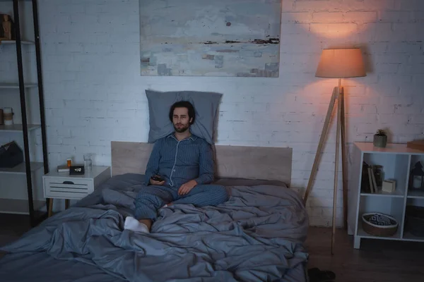 Homme avec insomnie tenant télécommande tout en étant assis sur le lit la nuit — Photo de stock