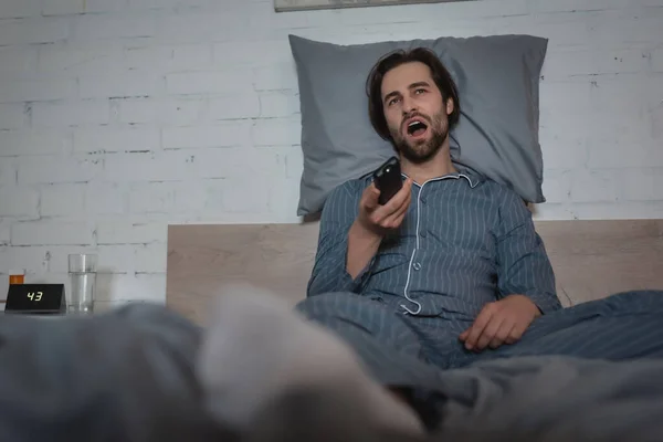 Homem com distúrbio do sono bocejando e segurando controlador remoto enquanto sentado na cama — Fotografia de Stock