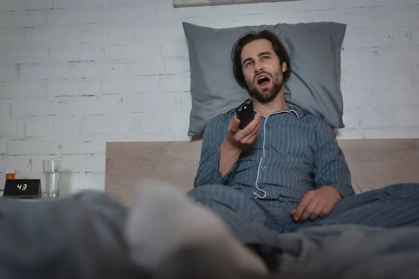 Homme avec trouble du sommeil bâillant et tenant télécommande tout en étant assis sur le lit — Photo de stock