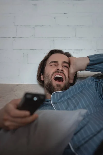 Tired man yawning and holding remote controller on bed at home — Stock Photo