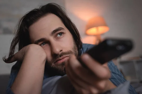 Brunette man with sleep disorder holding blurred remote controller in bedroom — Fotografia de Stock