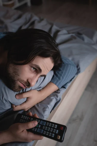 Schlafloser Mann hält Fernbedienung in der Hand, während er im Schlafzimmer auf Kissen liegt — Stockfoto