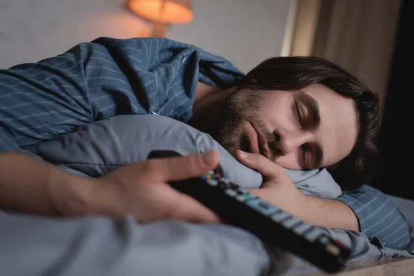 Jeune homme en pyjama tenant télécommande floue tout en dormant sur le lit à la maison — Photo de stock