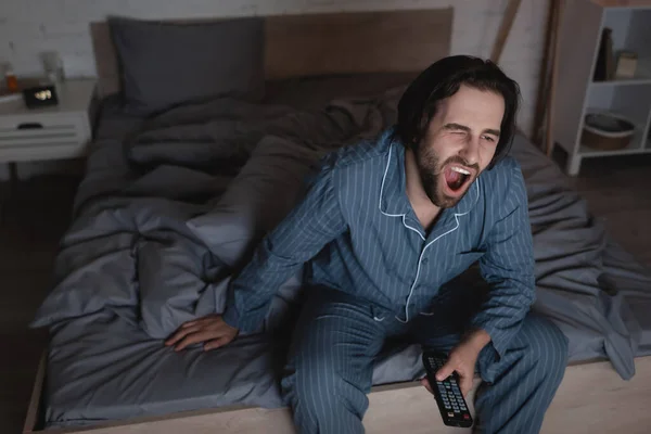 Уставший мужчина зевает и держит пульт дистанционного управления в спальне ночью — стоковое фото