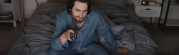 Homme insatisfait en pyjama en cliquant sur les canaux tout en étant assis sur le lit la nuit, bannière — Photo de stock