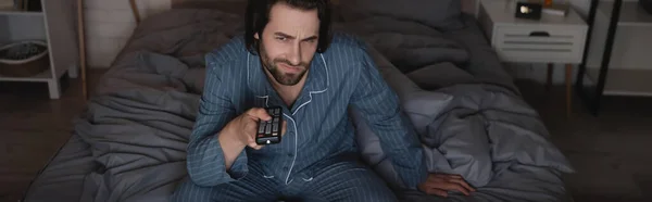 Dissatisfied man in pajama clicking channels while sitting on bed at night, banner — Fotografia de Stock