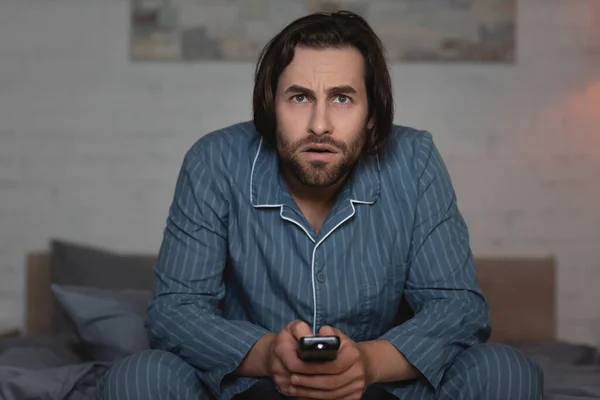 Man in pajama holding remote controller while watching tv on blurred bed — Stock Photo