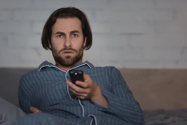 Нещасний чоловік у піжамі дивиться телевізор, сидячи на ліжку вночі — стокове фото