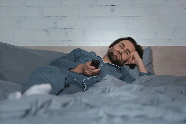 Tired man in pajama holding remote controller while lying on bed — Stockfoto