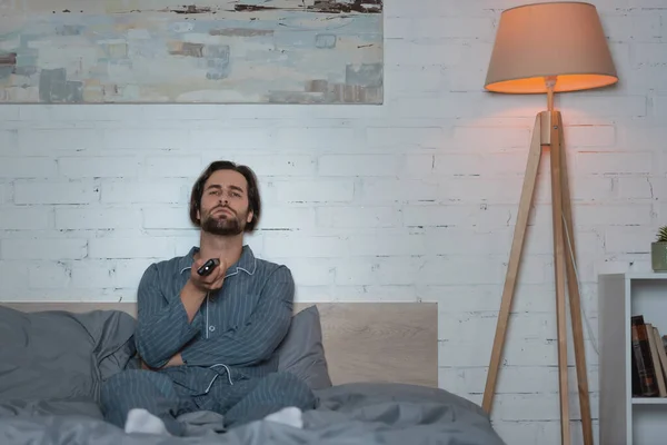 Bearded man with sleep disorder clicking channels while sitting on bed at night - foto de stock