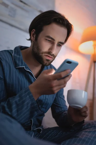 Young man holding cup of coffee and using smartphone in bedroom at night — Stock Photo