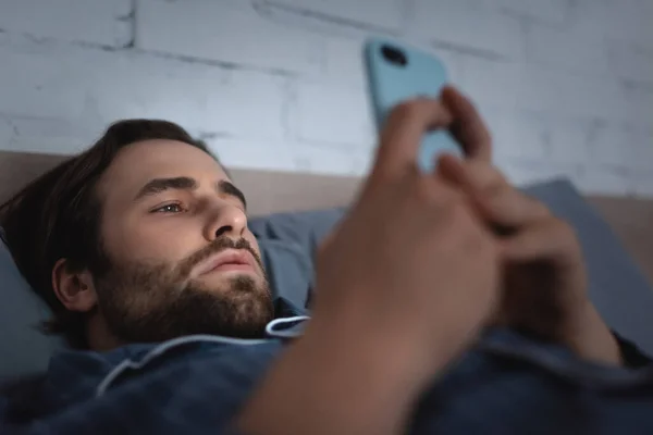 Bearded man in pajama using blurred smartphone on bed at night — Stock Photo