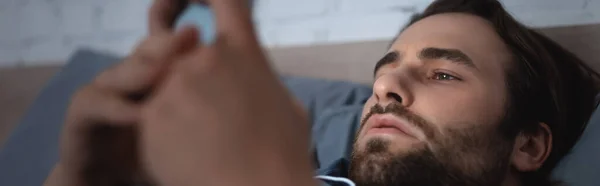 Бородатий чоловік використовує розмитий мобільний телефон у спальні, банер — стокове фото
