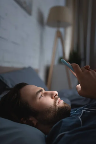 Side view of young man with sleep disorder using mobile phone on bed at night — Stock Photo