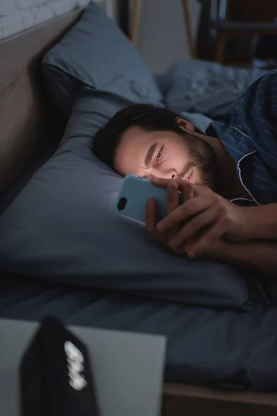 Sleepless man using mobile phone near blurred alarm clock on bed at night — Stock Photo