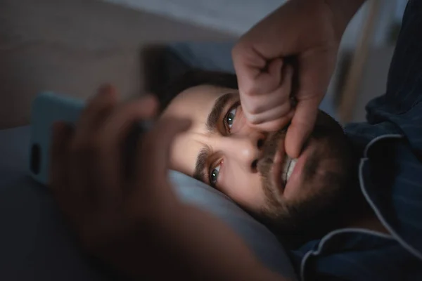 Наголошений чоловік кусає цвях на руці і використовує смартфон на ліжку вночі — стокове фото