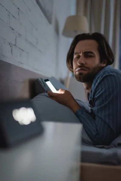 Uomo scontento utilizzando smartphone e guardando la sveglia offuscata sul letto di notte — Foto stock