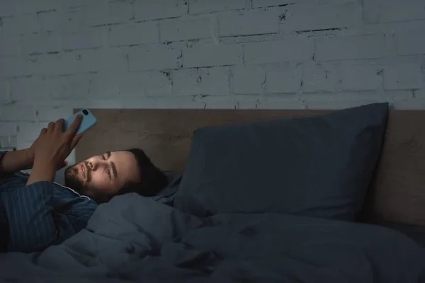 Young man using smartphone on bed at night — Stockfoto