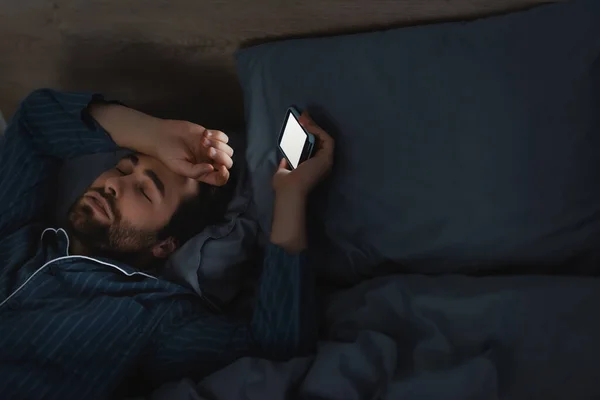 Hombre joven con trastorno del sueño sosteniendo el teléfono celular en la cama en el dormitorio - foto de stock