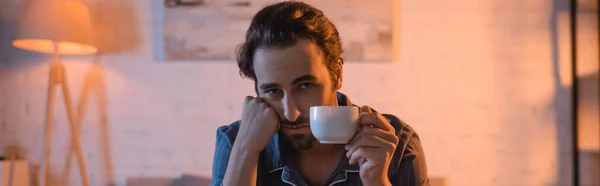 Hombre cansado en pijama sosteniendo la taza y mirando a la cámara en el dormitorio por la noche, pancarta - foto de stock