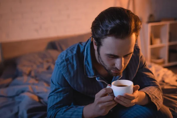 Junger Mann im Pyjama hält nachts Tasse Kaffee auf dem Bett — Stockfoto