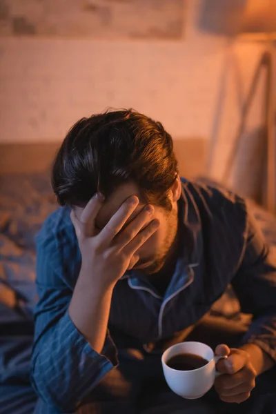 Tired young man holding cup of coffee while sitting on bed at night — Photo de stock