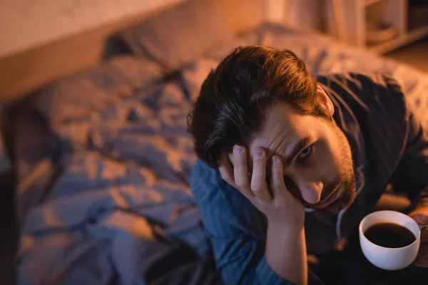 Erschöpfter junger Mann hält Tasse Kaffee in der Hand und blickt nachts in die Kamera auf dem Bett — Stockfoto