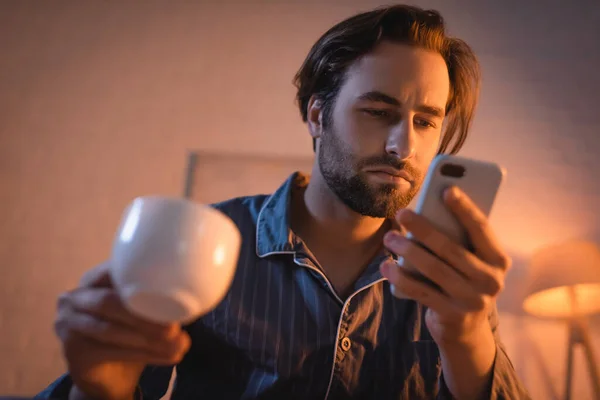 Joven en pijama sosteniendo café borroso y teléfono inteligente por la noche - foto de stock